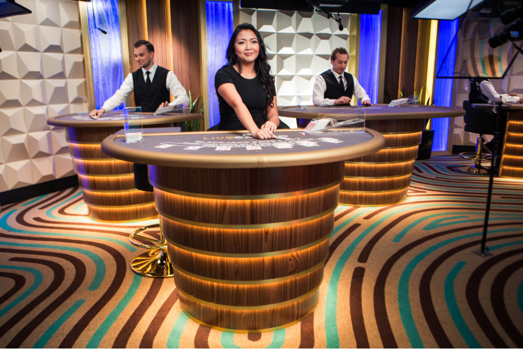 Evolution Gaming live dealer studio scene with three illuminated gaming tables and professionally dressed hosts standing behind them; warm gold and blue vertical lighting, textured geometric wall panels and a patterned carpet create a premium broadcast set. Foreground table shows a card game layout with chips and dealer area. Prominent messaging emphasizes customer support availability and responsible gaming features: 24/7 live chat, phone and email support, multilingual assistance, deposit/session limits, self-exclusion options and referrals to specialist services.