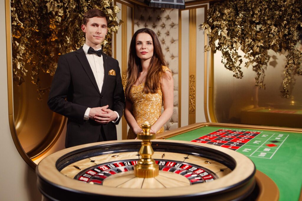 Evolution Gaming responsible gaming banner showing a formally dressed dealer in a tuxedo and a host in a gold dress standing beside a roulette wheel and green betting layout in an elegant, gold-accented studio; the image highlights safer-play messaging, deposit and session limits, self-exclusion options, and available multilingual support and referrals to specialist help.